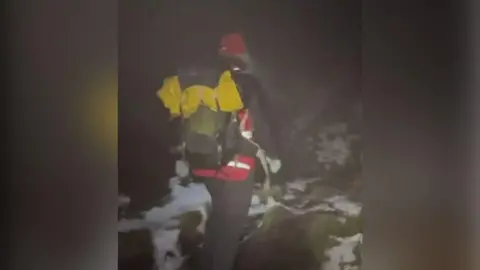 Mountain rescue team volunteer in climbing gear walking in wintry conditions with snow on the hills. He is pictured from behind. It is dark and raining. He has a red hi vis coat and a yellow equipment at the top of his green and grey ruck sack. 