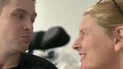 JO DUNNE Ethan and Jo looking into each other's eyes, both are smiling. He has short brown hair and she has blonde hair that is tied back and glasses perched on her head.