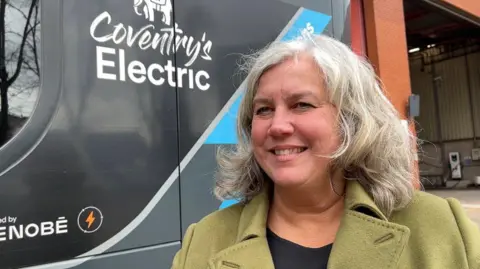 Heidi Alexander stands in front of an electric bus with the slogan "Coventry's Electric" on the side. She wears a green jacket.