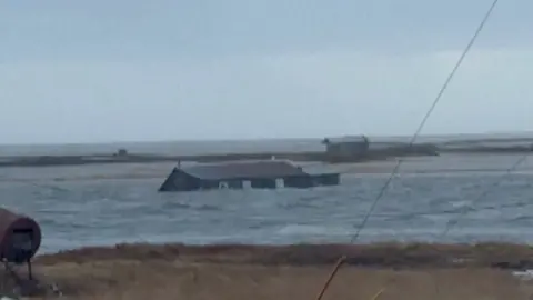 A house submerged almost up to its roof floats in water near land.