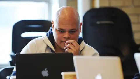 A photo of Conservative MP Jamil Jivani at a desk looking down at an Apple portable computer with a black cover. He has his left hand on his face, and is wearing a hooded sweatshirt that is dark blue and light grey. He is bald with tanned skin. 