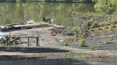 The amphitheatre has weeds growing out of the concrete with its wooden pontoon fenced off and damaged next to a pond in the park.