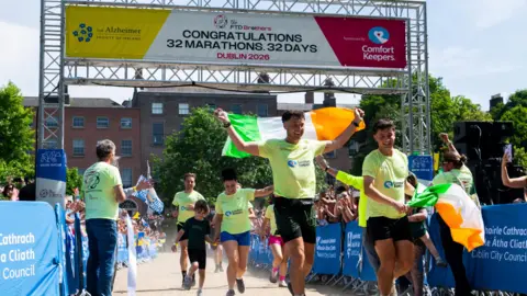 PA Media Several people, including a child, are seen racing across a finishing line. They are wearing green T-shirts, holding an Irish flag, while crowds watch on.