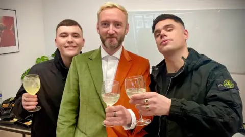 Kneecap trio posing for a photo - they are all holding a wine glass with some kind of light yellow coloured cocktail and a sprig of thyme. Mo Chara (who has short brown hair) is wearing a black zipped hoodie, DJ Provaí (who has bleached blonde hair and a light beard) is wearing a half green and half orange suit jacket with a white shirt, Móglaí Bap (has short black/brown hair) is wearing a dark green coat and t-shirt. 