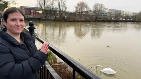 BBC News A smiling woman stands at the side of a large pool of water with swans nearby. 