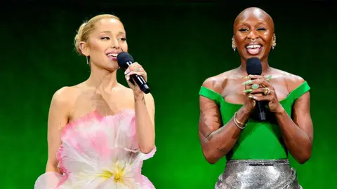 Getty Images Ariana Grande and Cynthia Erivo speaking on stage. The backdrop is green. Ariana is on the left, wearing a floral dress, speaking into a black microphone. Cynthia is wearing a green top and silver shiny skirt, smiling and speaking into a black microphone.