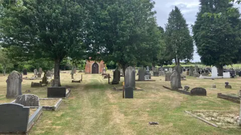 BBC/Lorna Bailey Graves and grass are in the foreground, trees are in the top half of the image and a building is in the background.