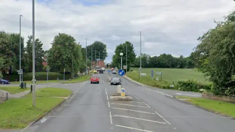 A61 junction with Jumbles Lane in Lofthouse, with the road fairly busy with vehicles. A black car is turning right, with a red car continuing along the A61.  There's grass and trees on both sides of the road. 