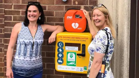 Clare Seed Two women, one with a black bob, wearing silver dangly earrings, a blue and white patterned top, and blue jeans, and another with shoulder length blonde hair, black glasses perched up on her head, a shoulder bag and a white and blue floral dress, smile as they rest their arms on a yellow defibrillator case.
