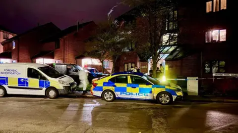 Leicester Media Online A number of police vehicles outside a block of flats at night. One van says FORENSIC INVESTIGATION. A forensic officer in white overalls is visible from the road. There is also police tape across the entrance way to the block.