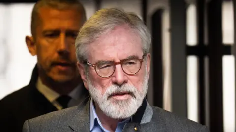 Gerry Adams has short grey hair and facial hair. He has round glasses and a grey suit. 