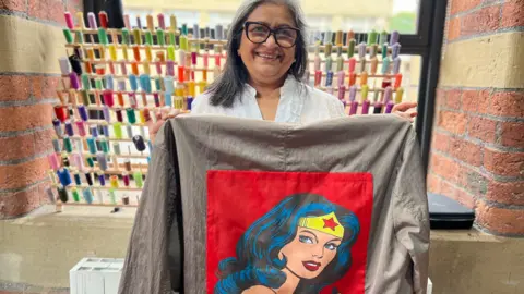 BBC/Aisha Iqbal A lady with shoulder-length black and grey hair and glasses holds up a grey jacket with a red Wonder Women logo on it
