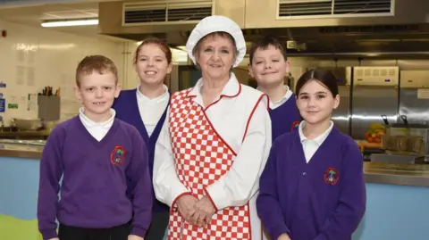 Gillian Morris in white overalls and catering hat and red and white checked apron with four pupils in canteen in uniform of purple jumper or cardigan and black trousers and white polo shirt