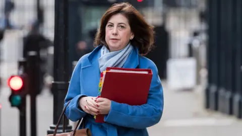 Lucy Powell seen wearing a blue jacket while holding a handbag and a red folder on the street.