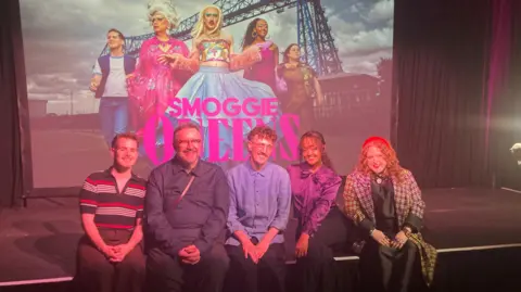 The cast of Smoggie Queens sit on a stage smiling at the camera in front of a big screen showing them dressed as their characters.