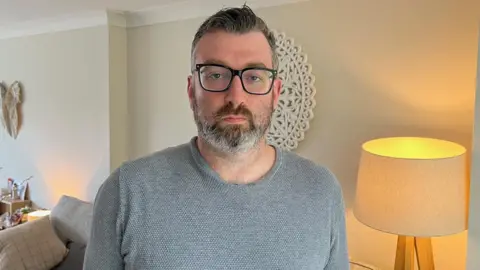Jamie Coulson/BBC A man in a grey jumper photographed in a living room, with a sofa in the background and a lamp to one side. He wears a grey jumper and glasses.