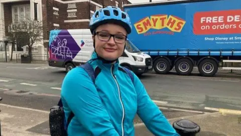 Sally Fairfax/BBC A woman in a bright blue fleece wearing a backpack and a blue helmet. She has rectangular glasses and is smiling at the camera. Her arms appear to be reached out holding a bike. A road is behind her with a few vehicles driving on it.