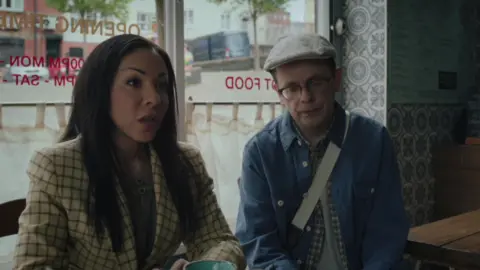 Two characters - one woman and one man - sitting in a cafe in conversation with an unseen figure on the other side of the table. The woman is wearing a yellow and brown check jacket and the man is wearing a denim jacket, glasses and a white cap