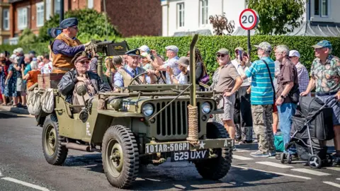 Andy Bell Images A vintage military jeep drives along a road during a wartime-themed parade. Several people in period military uniforms ride in the vehicle, while crowds of spectators line the pavement behind them.