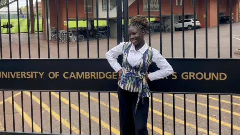Francisca Arhinful Francisca Arhinful standing in front of a black metal gate which has University of Cambridge written across it. She is smiling broadly, with her hands on her hips. Beyond the gate is a red brick building and parked cars and bikes. 