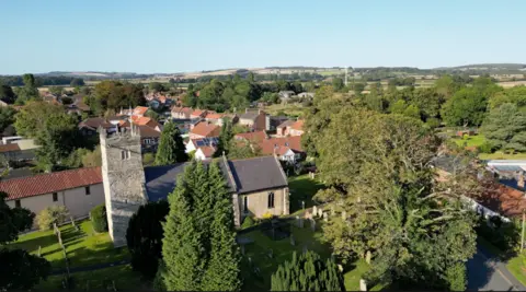 A bird's eye view of Shiptonthorpe, lush green, tall trees cast shadows over the village church. In the distance houses are nestled in between greenery.