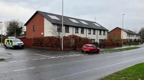 BBC a police van and a red car are parked on the road, with houses behind it