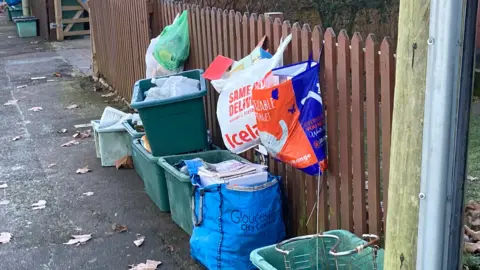 Green recycling boxes and a blue bag full of paper to be recycled are left outside a brown garden fence to be collected. There are several carrier bags, also full of recycling, hung upon the fence.