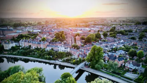 An aerial view of Shrewsbury, in the bottom of the frame is a river with a footbridge suspended above it. Next to the river is a long stretch of road lined with trees and other greenery. Behind that are several streets of houses and other buildings, mostly made of red brick