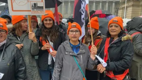 A group of women are wearing orange hats with the GMB Union logo on them. They are holding banners and flags that call for equal pay. They are wearing coats and jackets, and people in the crowd behind them are holding umbrellas.