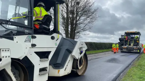 Laura Foster/BBC A white steam roller on top of newly laid tarmac. Another steam roller is further along the road. A number of people in high visibility clothes trousers and jackets are around. 
