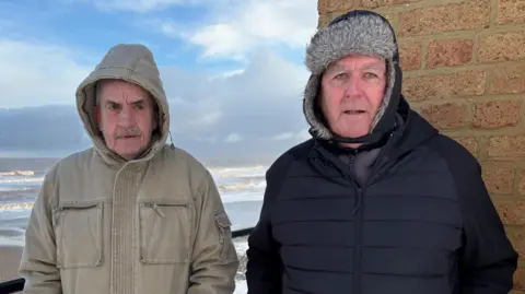 Kevin Shoesmith/BBC Two men, both in their 60s, are standing with a stormy sea behind them. Both have their hoods up to afford them protection from the wind.