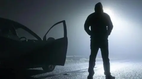 Getty Images A man in silhouette stands in the middle of the road beside a car. 
The car door is open and it looks like he is waiting for someone. 