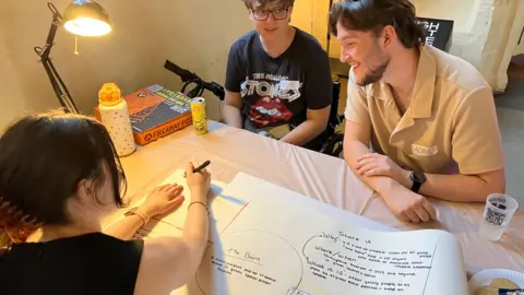 Jon Wright/BBC Three young people sit round a table with a large sheet of paper in the middle with circles and writing on.