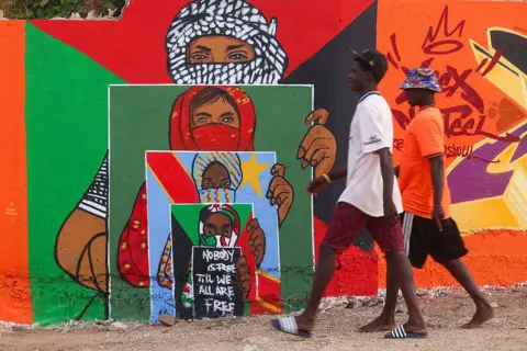 CEM OZDEL / ANADOLU / GETTY IMAGES Men walk past a mural depicting people with the flags of Sudan, the Democratic Republic of Congo and the Palestinian territories.