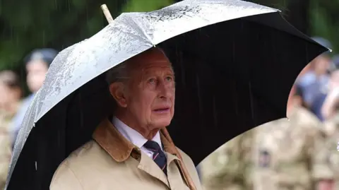 PA Media King Charles wearing a brown coat and holding a black umbrella standing in the rain.