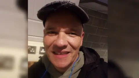 Lisa Lailey James Starr smiling at the camera. He has a dark brown cap on with green hoodie and black jacket. In the background are kitchen tiles. 