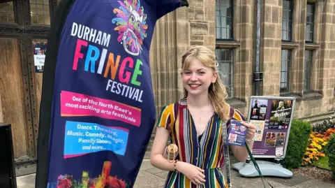 Isabel Maria is smiling at the camera at Durham's Palace Green last July. She is standing next to a navy blue Durham Fringe banner. She is wearing a blue, yellow, green and white striped dress. Her wavy blonde hair is half-up. 
