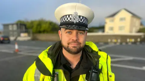 Sgt Elliot Butler looks at the camera, he wears a high-vis jacket and a white hat with the force's emblem on it. His jacket has a radio attached to it.