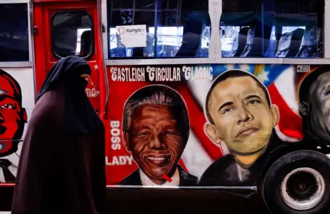 LUC GNAGO / REUTERS A woman wearing an abaya and niqab walks past a bus with artwork depicting late South African President Nelson Mandela and former US President Barack Obama, whose later father was Kenyan. 