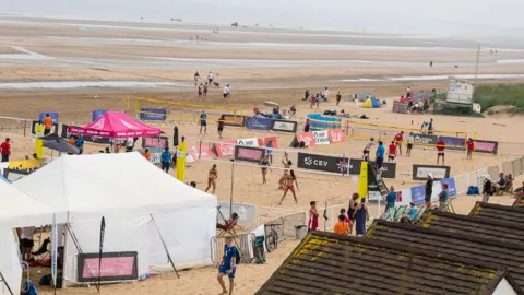 Children and young people can be seen on Bridlington beach playing on volleyball courts. The sandy beach stretches out into the distance. 