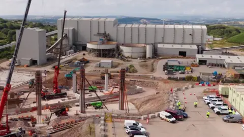 There are grey industrial buildings and huge upright structures at the mine. There are cars parked nearby and containers on the edge of the shot, with the moors in the background.