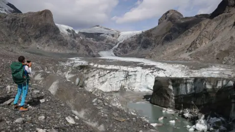 A scientist takes a photo as part of an annual monitoring project of Austrian glaciers administered by the Austrian Alpine Association in 2023. In the background the glacier slopes down the rocky mountainside, while in the foreground chunks of ice are in a stream.