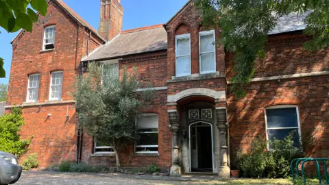 Clare Worden/BBC A large two-storey brick building with ornate stone columns around the entrance door. 