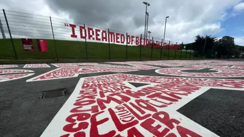 Large white writing on grey tarmac has red writing inside of it. The words of the names of Manx school children. Behind it the quote "I dreamed of being like my heroes" can be read in large red writing attached to a green fence by a grass hill.