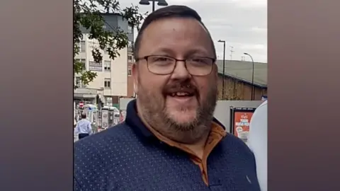 Hertfordshire Constabulary A smiling man, wearing glasses and a beard, is stood on a high street