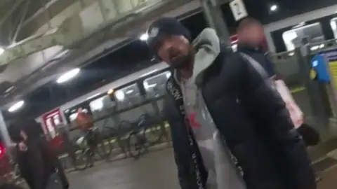 A still taken from the bodyworn police footage of Ashley Warren's arrest. He is walking along a platform with a train docked behind him. He is wearing a black beanie hat, black coat and grey hoodie.