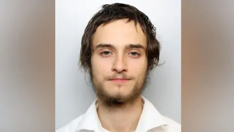 A young man with a thin beard and medium-length hair stares forward for a police mugshot. He is wearing a white shirt. 