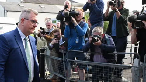 Getty Images Jeffrey Donaldson, a man with short grey hair and glasses is walking into a courthouse. Behind him are a bank of photographers taking his picture.
He is wearing a dark blue suit, a white shirt and green tie.