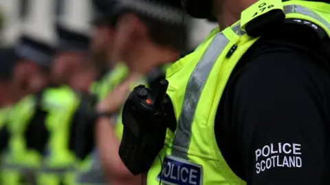 A close-up of a Police Scotland officer's upper torso showing a close-up of his radio, which is attached to his hi-vis vest. He is also wearing a short-sleeves black top with the Police Scotland logo visible on the left sleeve