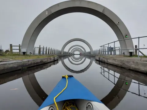 Raymond Carstairs It's a very cloudy and misty day. A view of the Falkirk wheel from a kayak on the waterway. You can see one end of the kayak and five steel arches over the calm waterway.